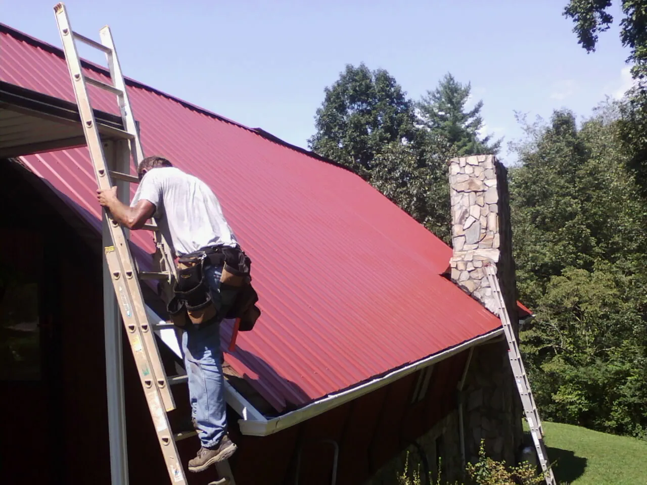Roofer on a ladder beside a red metal roof and stone chimney — Soffit & Fascia Repair by Roofing Brothers in Avenel